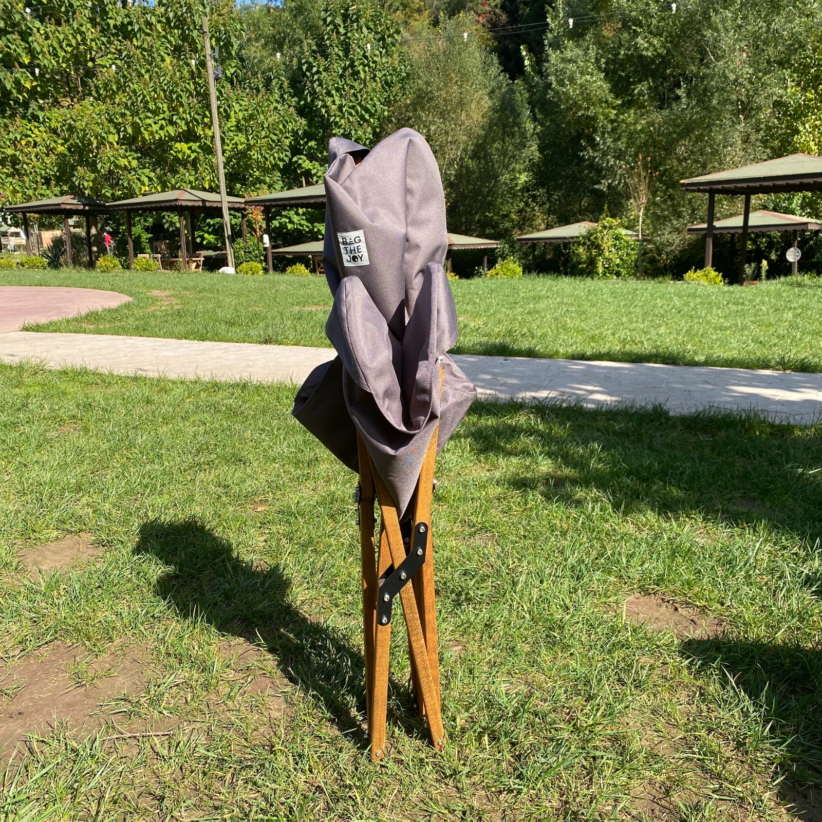 Gri bir katlanır sandalye parkta çimenlerin üzerinde duruyor. Arka planda ağaçlar ve piknik masaları var. Sandalye gölge düşürüyor.