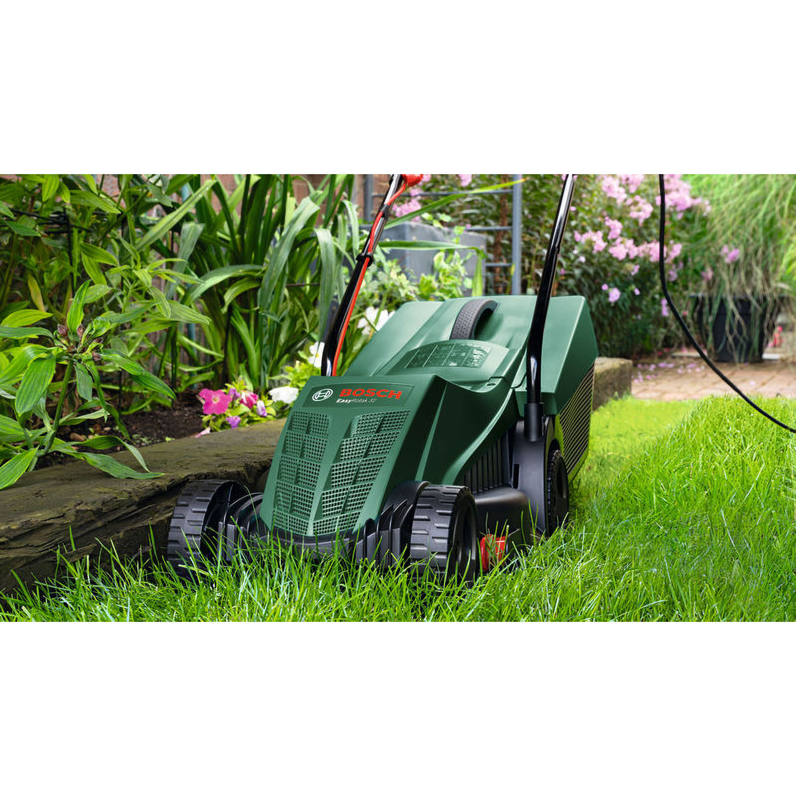 Ein grüner Rasenmäher in einem Garten. Der Mäher steht auf Gras, im Hintergrund Pflanzen und Blumen.