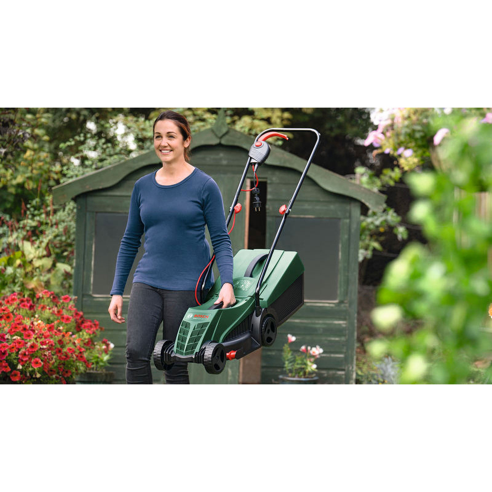 Eine Frau hält einen grünen Rasenmäher in einem Garten. Im Hintergrund befindet sich ein Schuppen, davor Blumen. Die Frau lächelt.