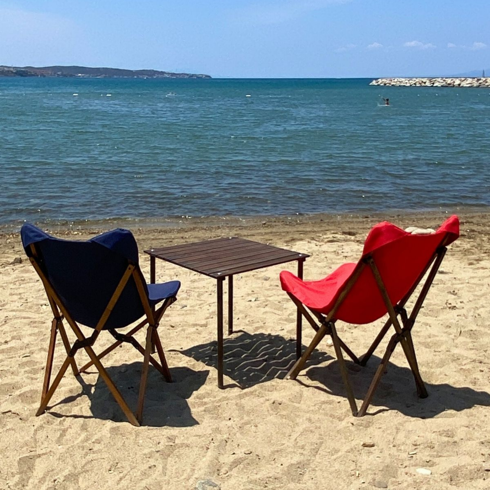 İki sandalye ve bir masa ile plaj sahnesi. Arka planda mavi deniz ve gökyüzü. Kumda kırmızı ve mavi sandalyeler.