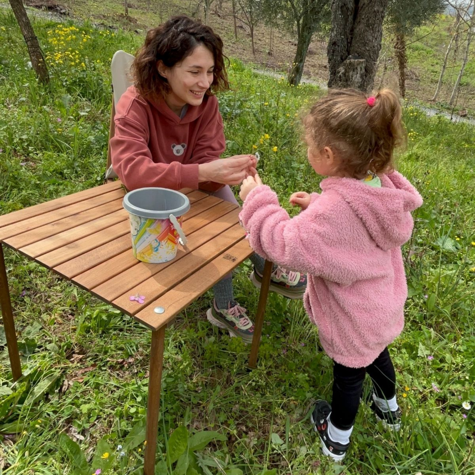 Bir kadın ve bir çocuk çayırda çiçeklerle oynuyor. Yakınlarda ahşap bir masa ve bir kova var.