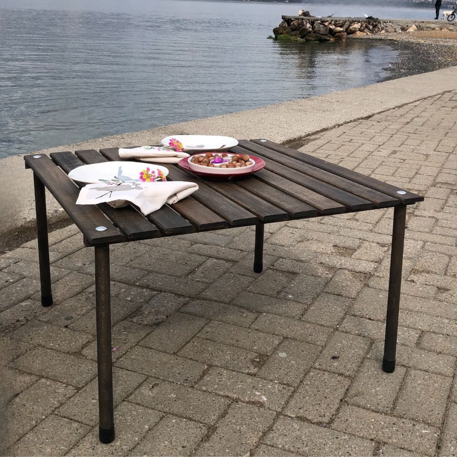 Su kenarında tabak ve kuruyemişlerle dolu masa. Çiçekli peçeteler. Ön planda taş kaldırım. Arka planda sakin deniz.