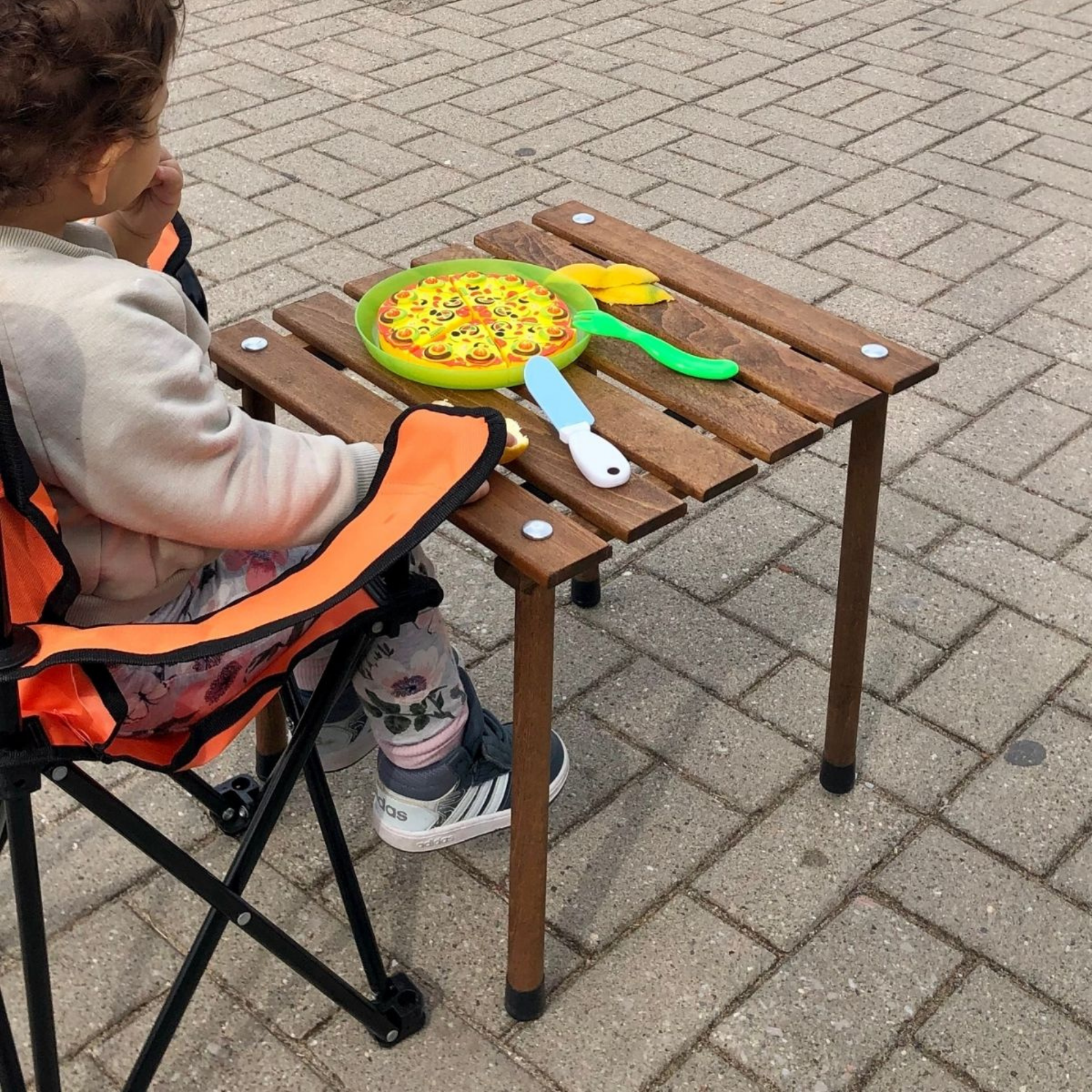Bir çocuk, üzerinde oyuncak pizza, çatal ve bıçak bulunan küçük bir ahşap masada oturuyor. Çocuk bir sandalyede oturuyor.
