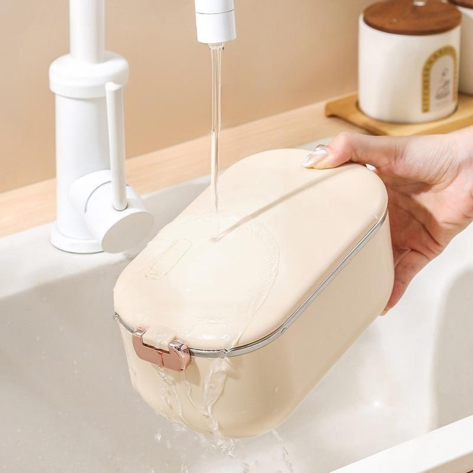 Eine beige Brotdose wird unter einem Wasserhahn gewaschen. Eine Hand hält die Dose. Wasser strömt aus dem Wasserhahn.