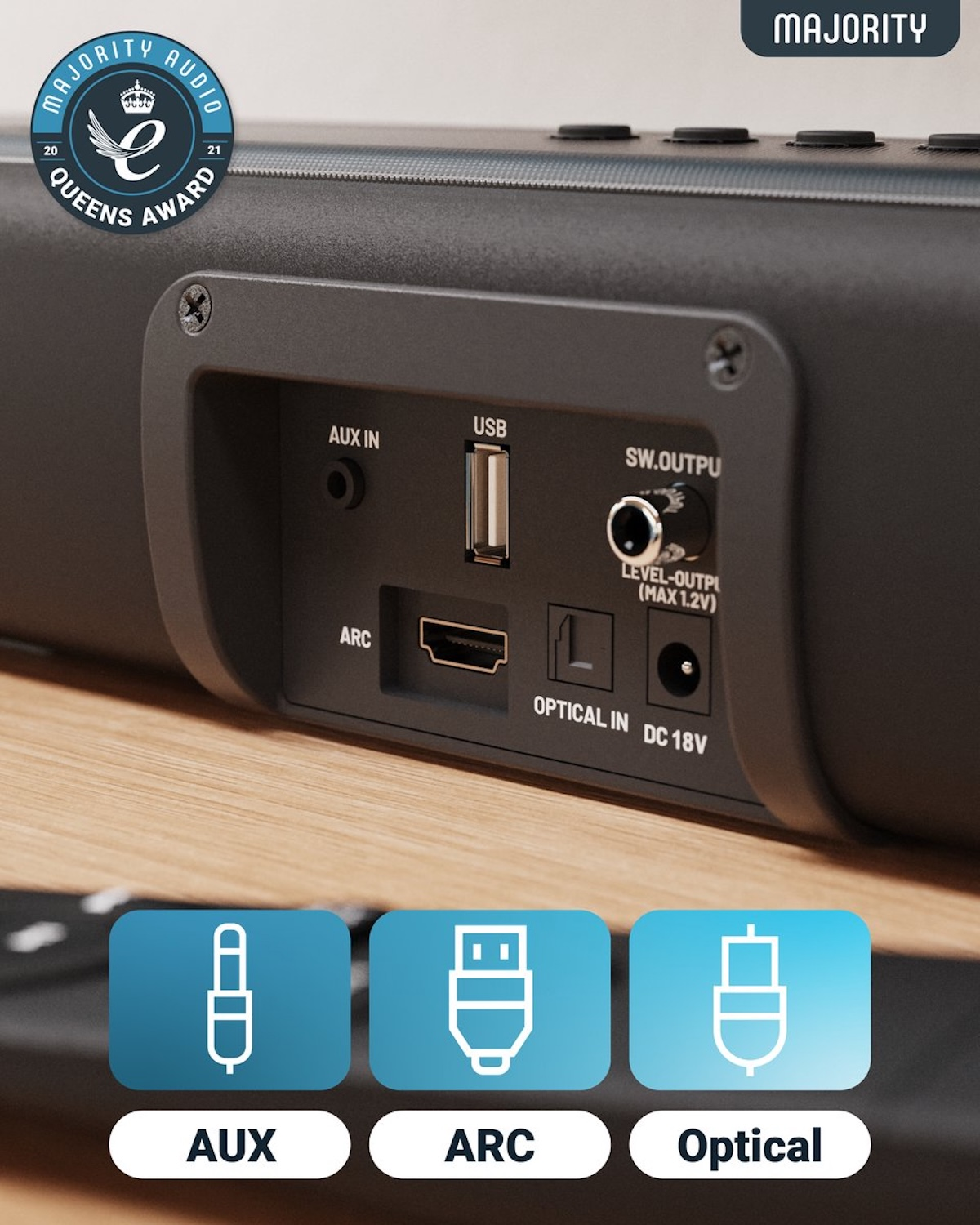 Black soundbar with ports: AUX, USB, ARC, Optical. Includes a Queens Award badge and Majority logo. On a wooden surface.