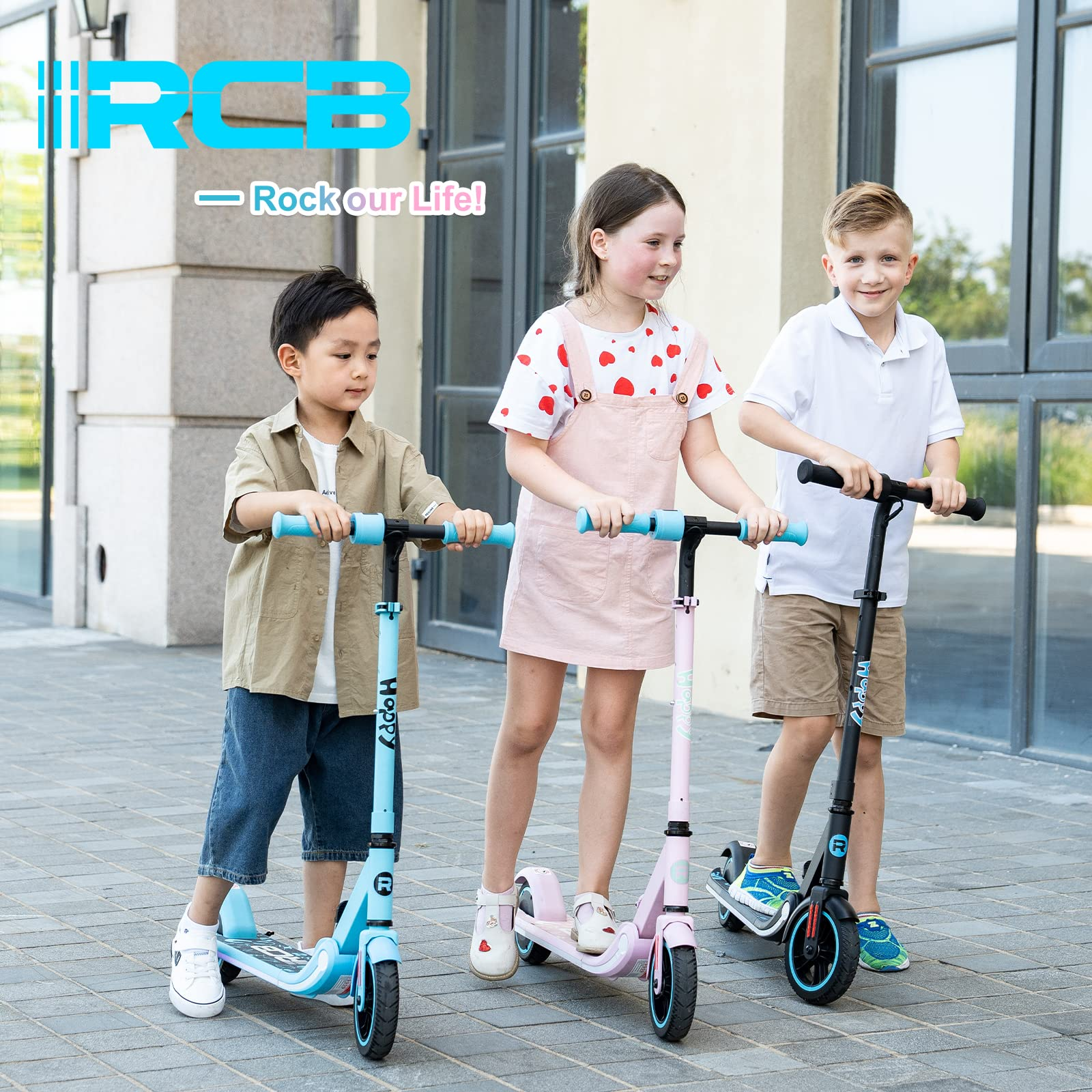 Three children with scooters. One blue, one pink, and one black. They are smiling and standing on a paved surface.