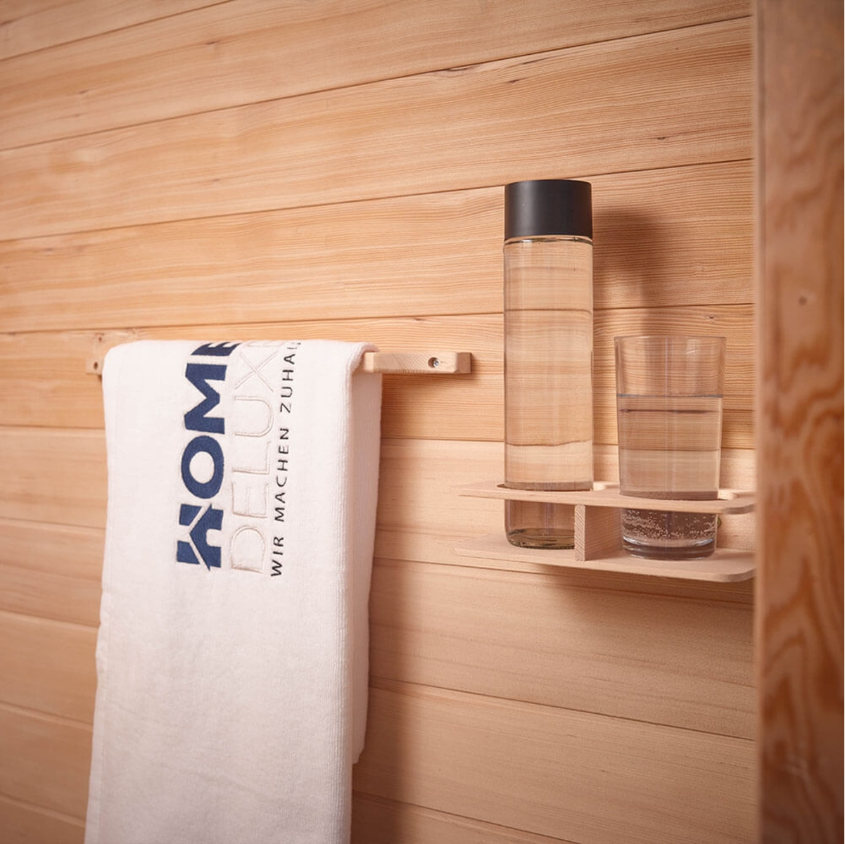 Ein hölzernes Sauna-Interieur mit einem Handtuch, einer Wasserflasche und einem Glas auf einem kleinen Regal. Die Wand ist aus Holz.