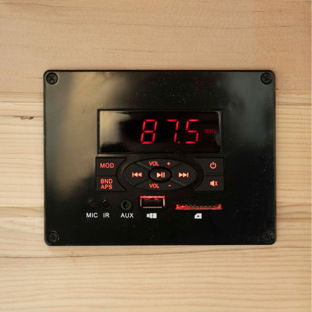 Schwarzes Bedienfeld mit roter Digitalanzeige, die 87,5 MHz anzeigt. Tasten für Lautstärke, Modus und Strom. Auf hellem Holz montiert.