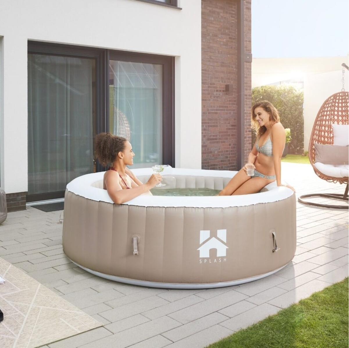 Zwei Frauen in Badeanzügen entspannen sich in einem aufblasbaren Whirlpool. Eine hält ein Getränk. Terrasse, Haus und Hängesessel im Hintergrund.