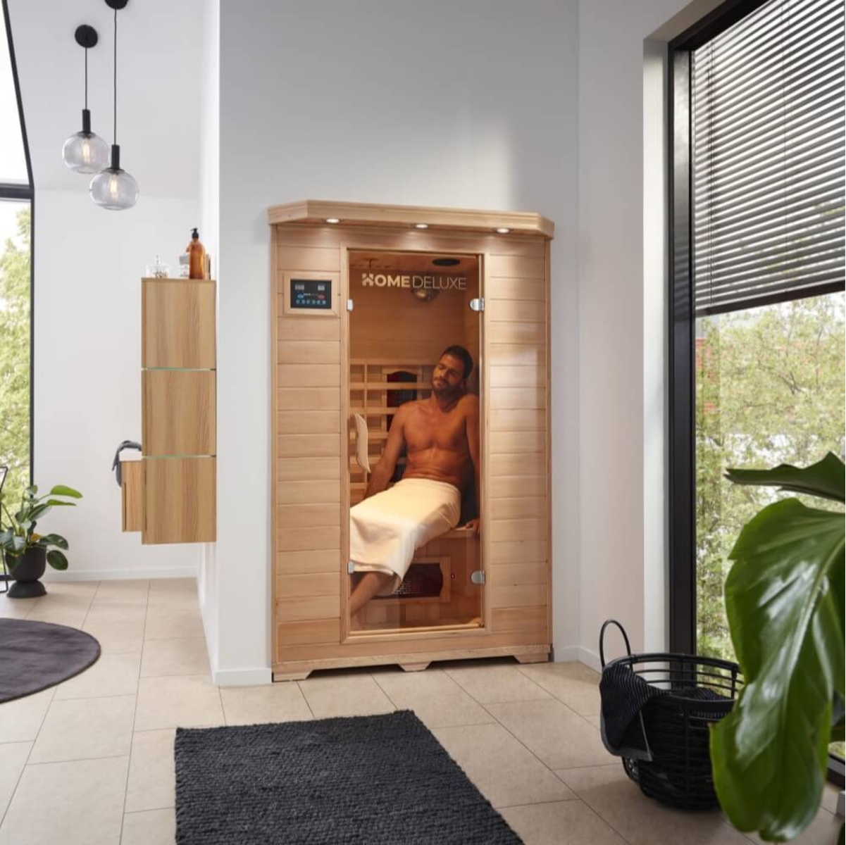 Mann in Sauna, in ein Handtuch gewickelt. Holzsauna mit Glastür. Raum mit Fenster, Pflanzen und schwarzem Teppich.