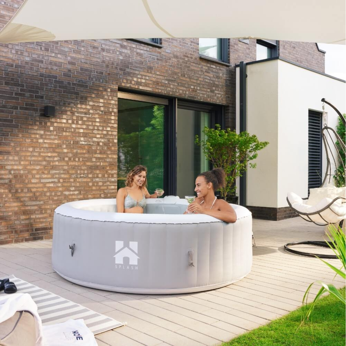 Zwei Frauen in einem grauen Whirlpool, trinken Cocktails, auf einer Terrasse. Backsteingebäude im Hintergrund, Hängestuhl und Rasen.