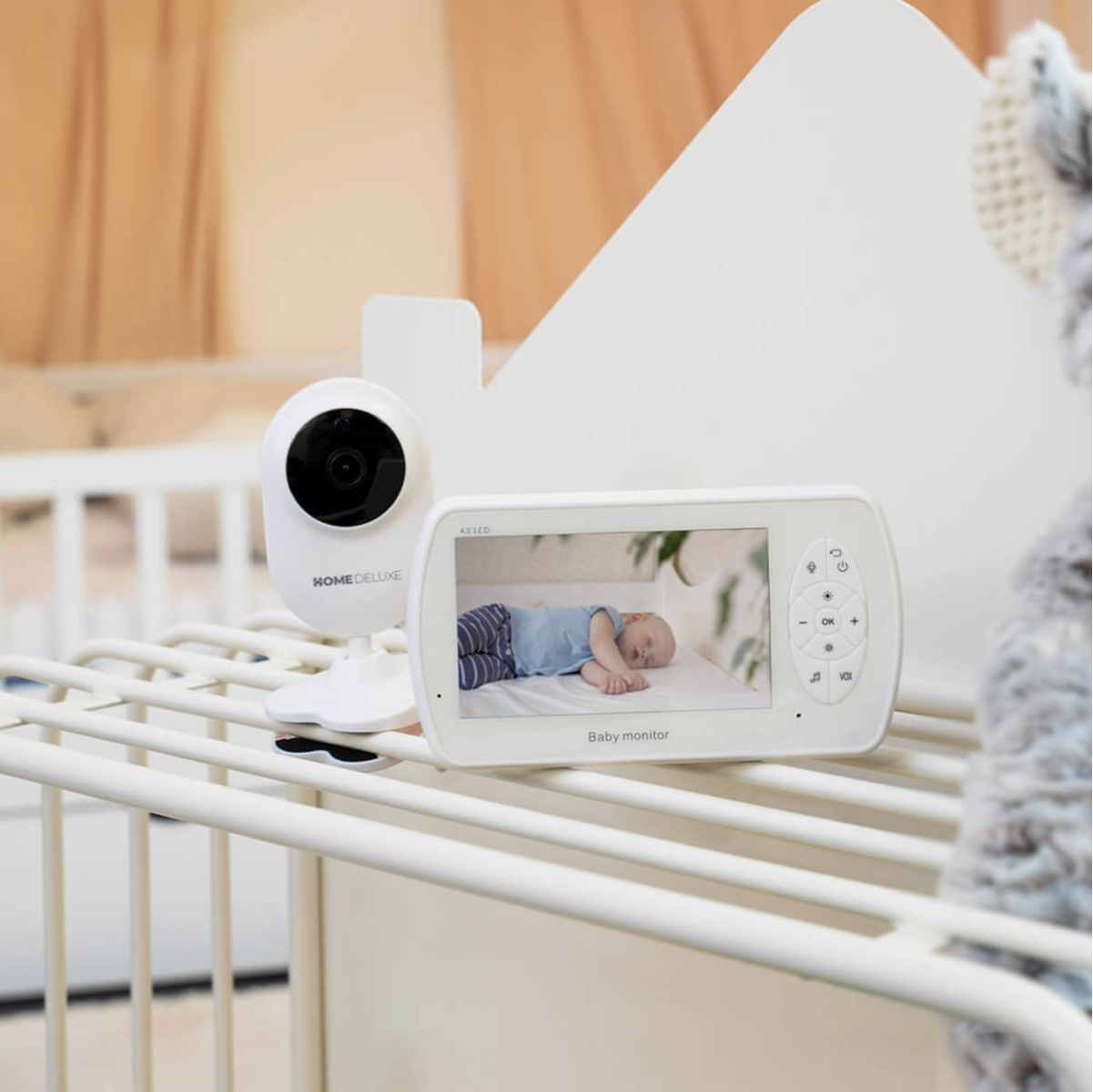 Ein Babyphone zeigt ein schlafendes Baby. Eine weiße Kamera und ein Monitor stehen auf einem weißen Kinderbett. Ein Stofftier ist rechts.