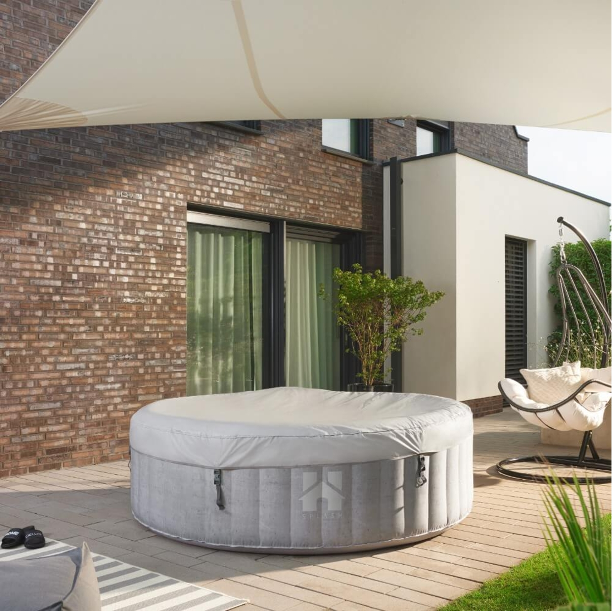 Ein runder Whirlpool mit Abdeckung steht auf einer Terrasse in der Nähe einer Backsteinmauer und einem weißen Gebäude.