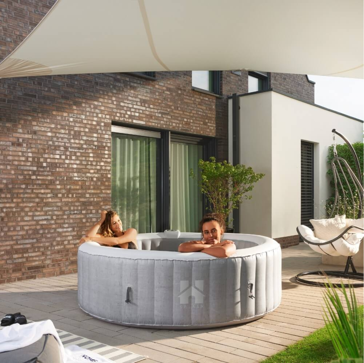Zwei Frauen entspannen sich in einem grauen aufblasbaren Whirlpool auf einer Terrasse. Eine Backsteinmauer und ein weißes Gebäude sind im Hintergrund.