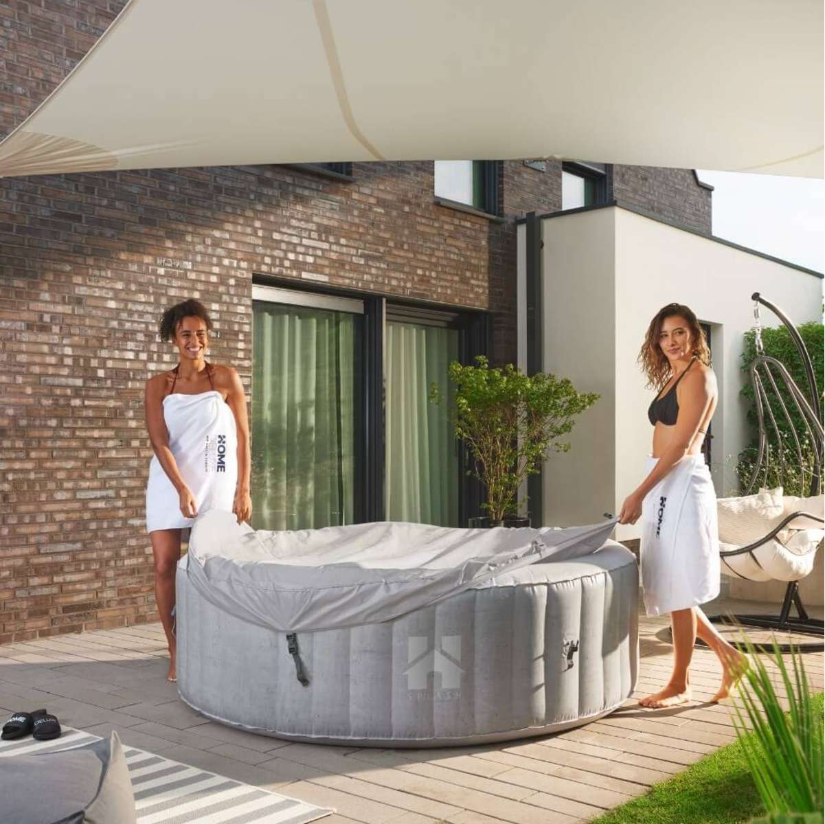 Zwei Frauen in Handtüchern decken einen Whirlpool auf einer Terrasse auf. Im Hintergrund eine Backsteinmauer und ein modernes Haus.