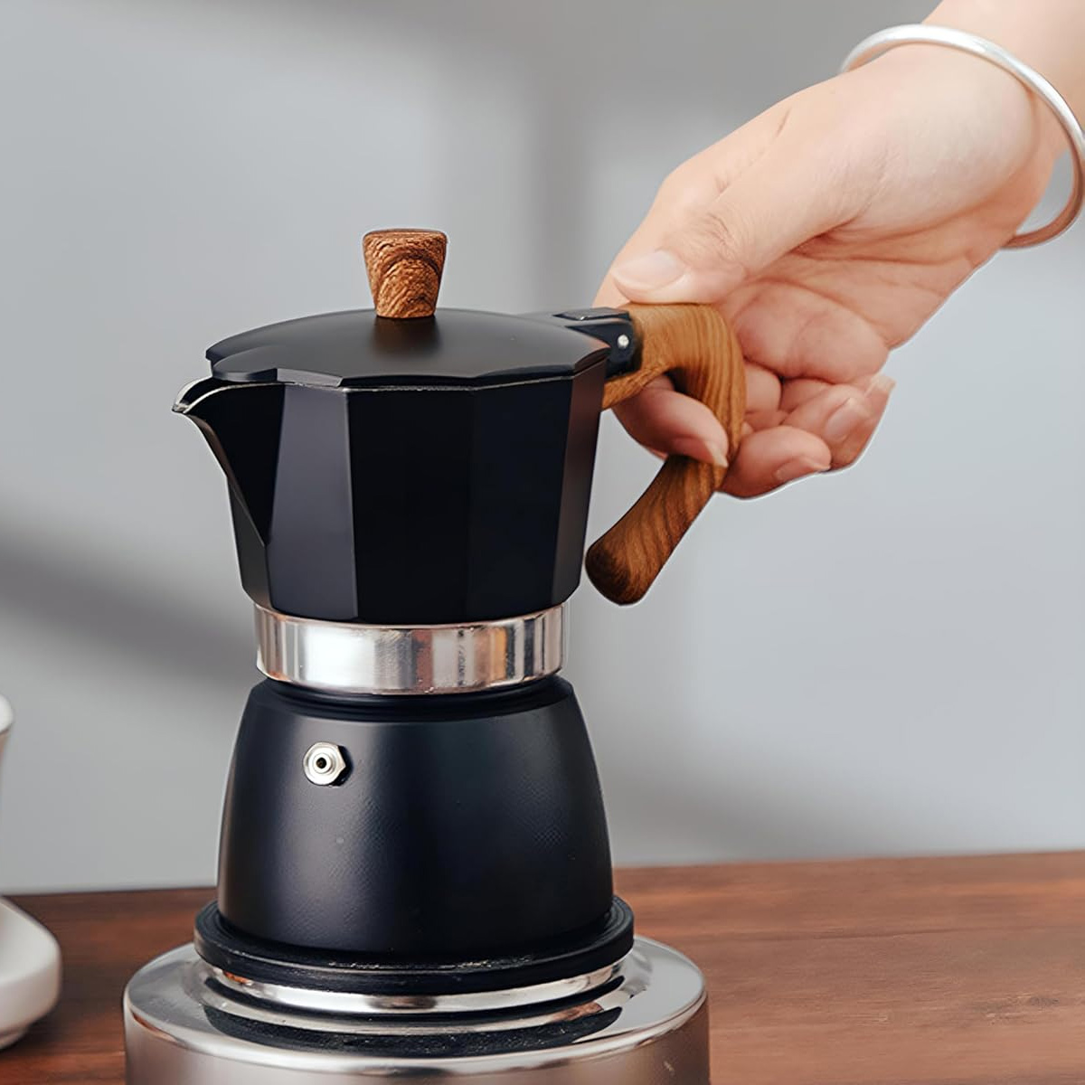 Eine Hand hält eine schwarze Espressomaschine mit Holzgriff. Die Espressomaschine steht auf einer silbernen Herdplatte auf einer Holzoberfläche.