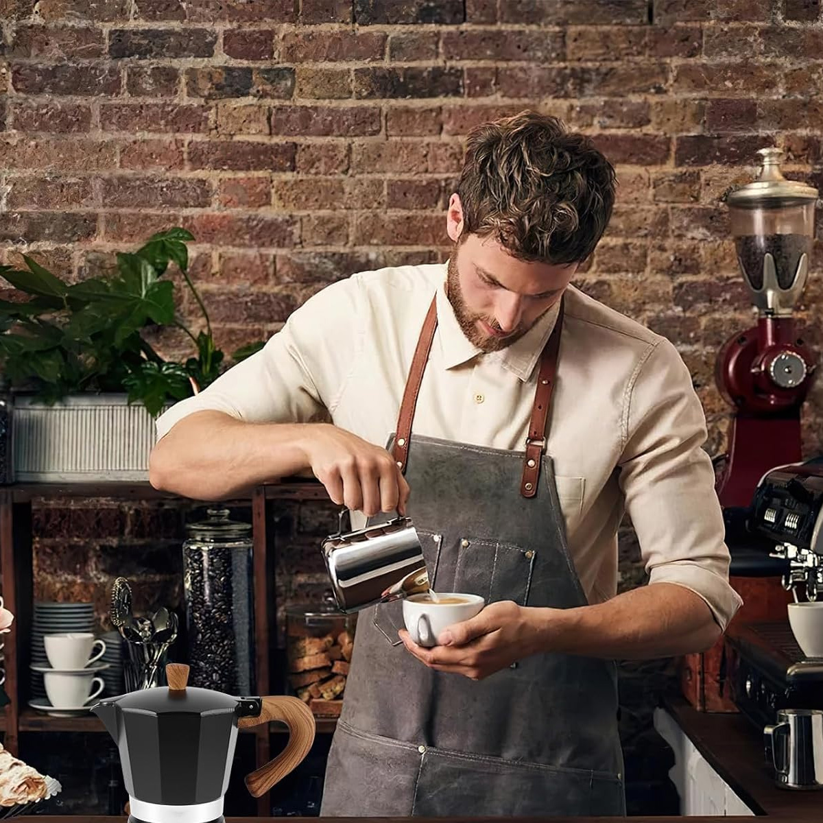 Ein Barista gießt Milch in eine Tasse Kaffee. Im Vordergrund befindet sich eine schwarze Kaffeemaschine. Der Hintergrund hat Backstein.