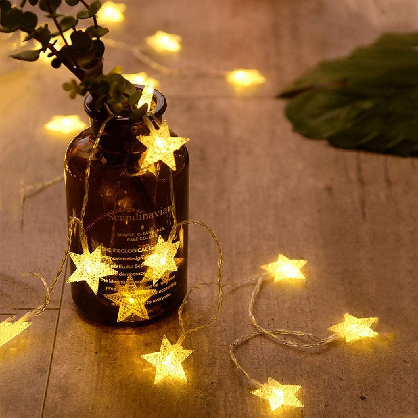Eine Glasflasche mit einer sternförmigen Lichterkette. Grüne Blätter befinden sich in der Flasche. Mehr Sternlichter liegen auf dem Tisch.