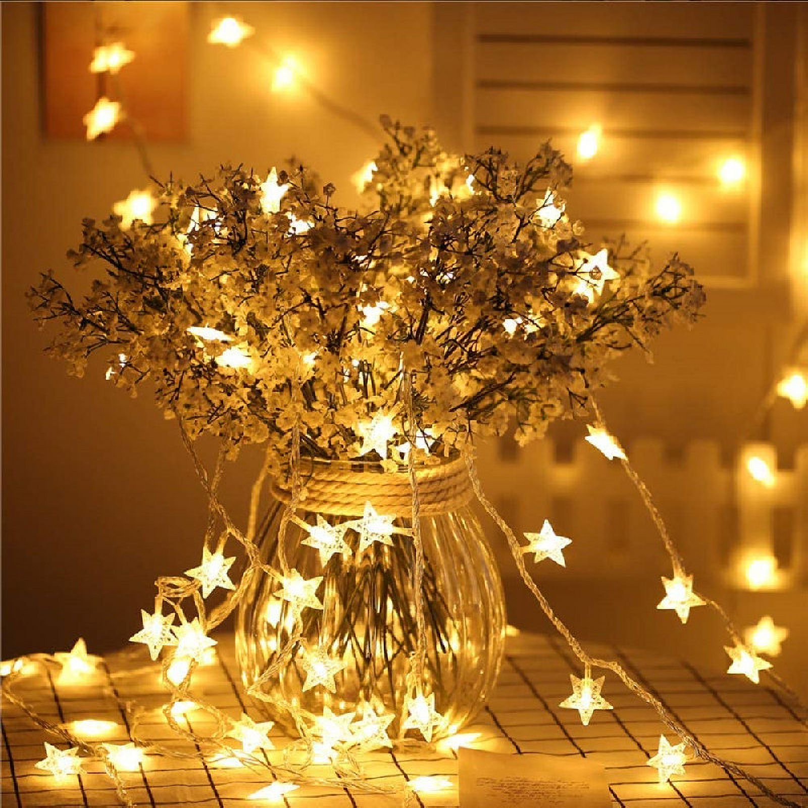Eine Glasvase enthält Blumen und sternförmige Lichter. Schnüre mit Sternlichtern drapieren sich um die Vase und auf einer karierten Oberfläche.