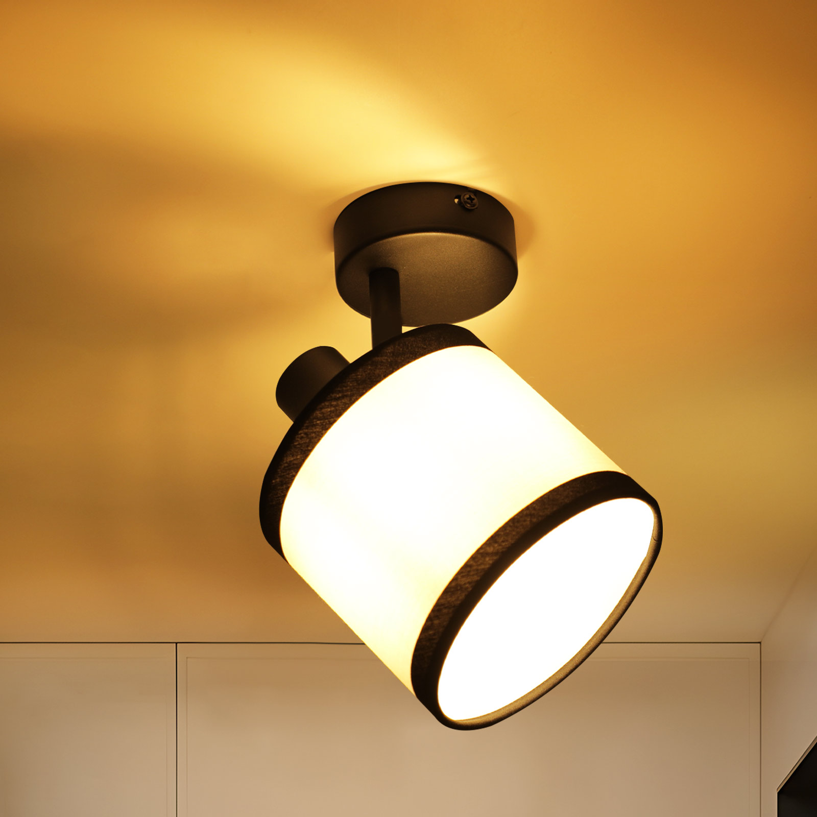 Un faretto da soffitto con paralume bianco. Accenti neri sui bordi del paralume. Illuminazione calda. Il soffitto è beige.