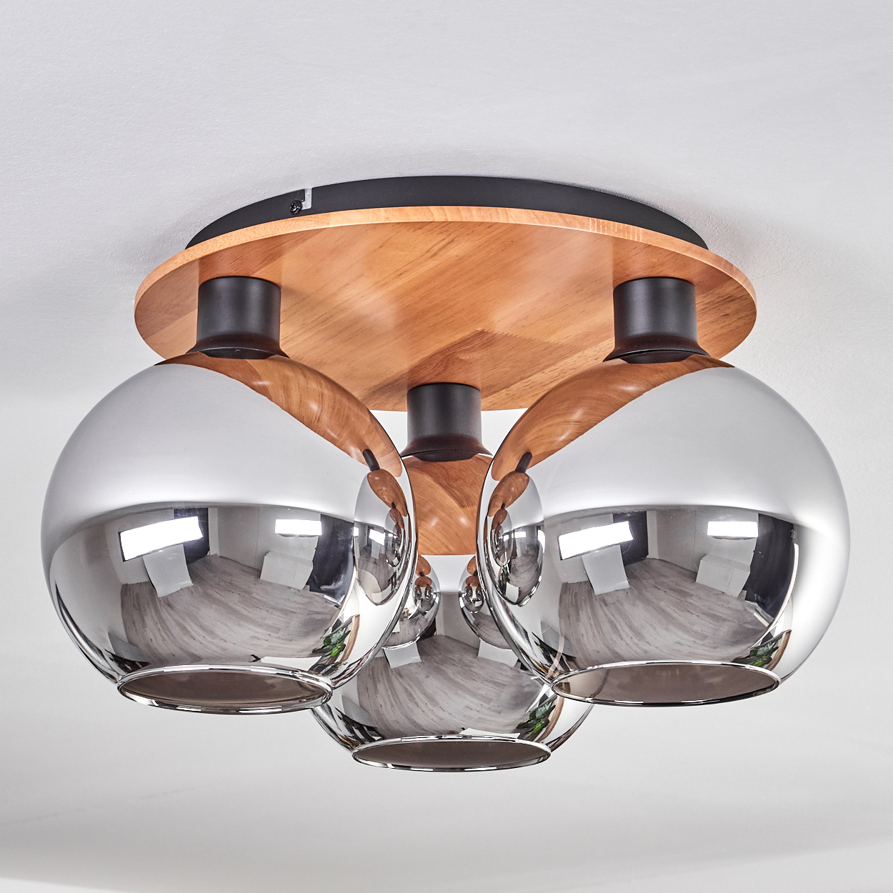 Eine Deckenlampe mit drei kugelförmigen Chromschirmen. Sie hat einen Holzsockel mit schwarzen Akzenten, vor einer weißen Decke.