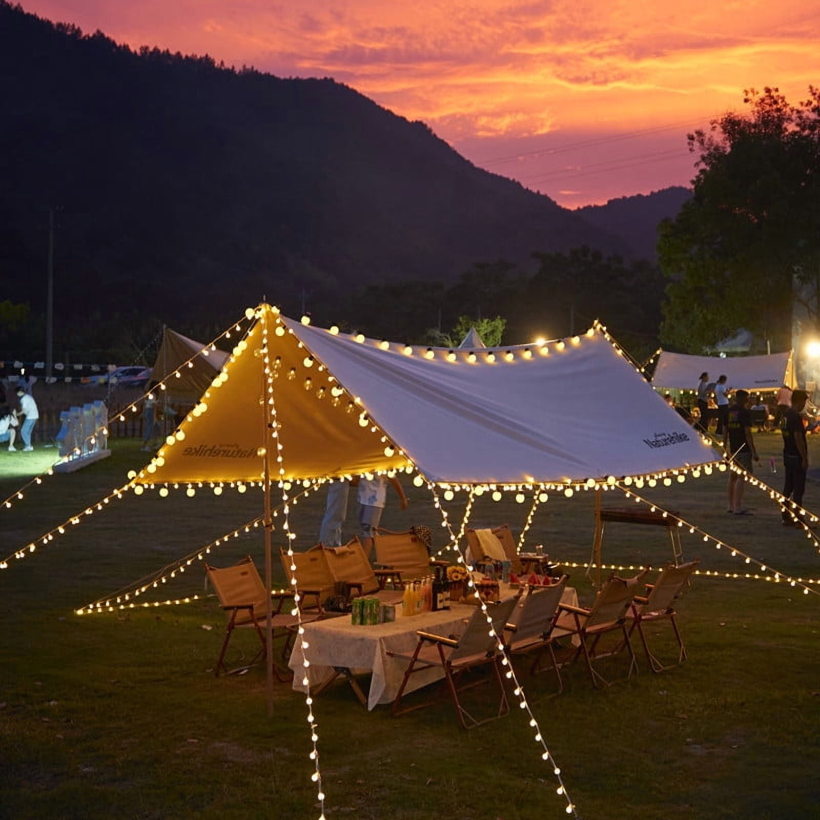 Ein Campingplatz in der Dämmerung. Ein Zelt mit Lichtern über einem Tisch und Stühlen. Im Hintergrund sind Menschen, am Himmel ein Sonnenuntergang.