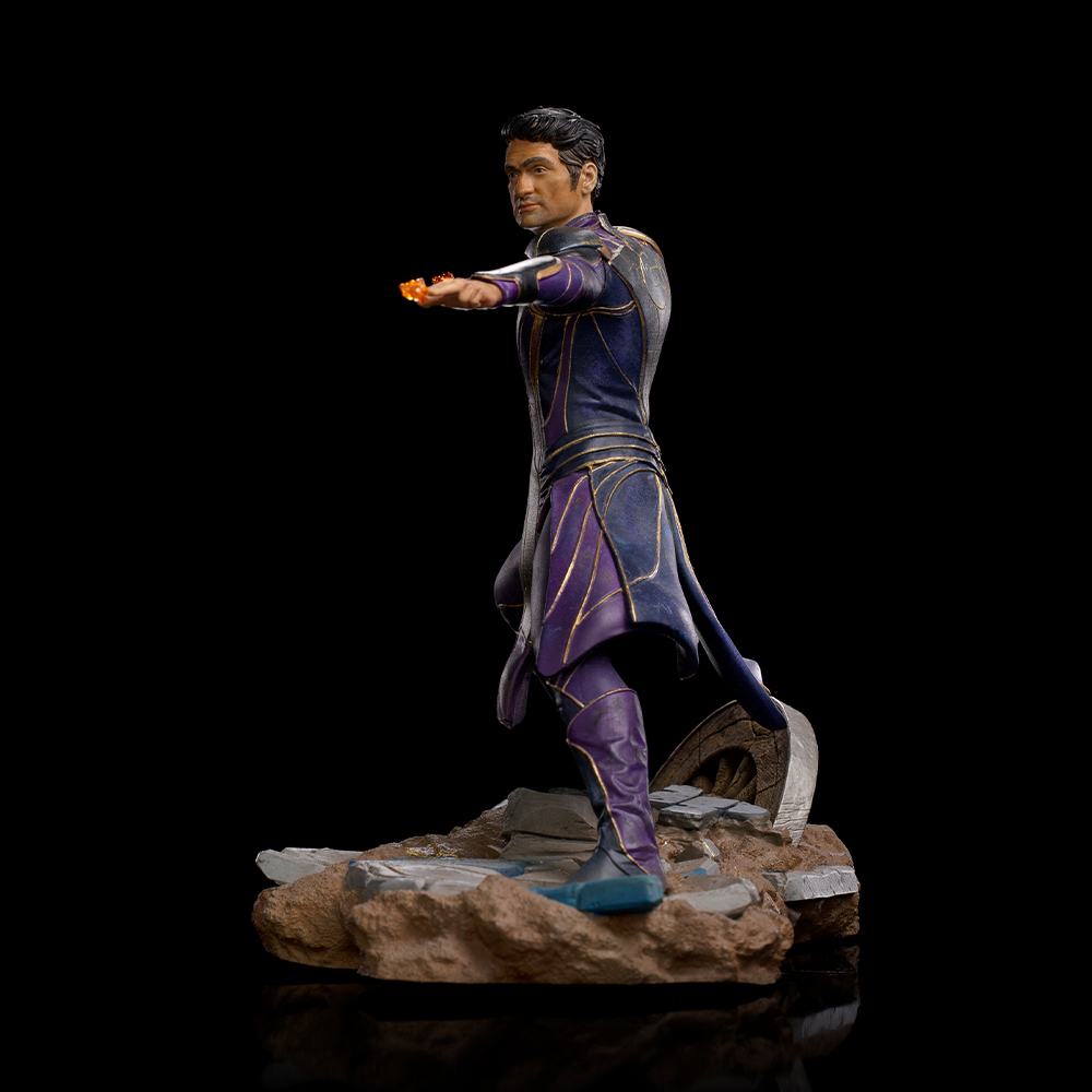 Una statua di un uomo in costume blu e viola, con in mano un oggetto arancione brillante. È in piedi su rocce con sfondo scuro.