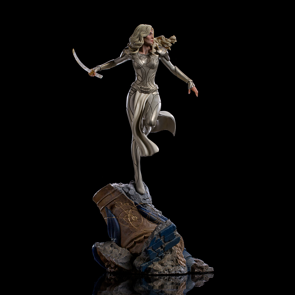 Statua di donna bionda, in abito argento, con lama, su roccia, sfondo nero.