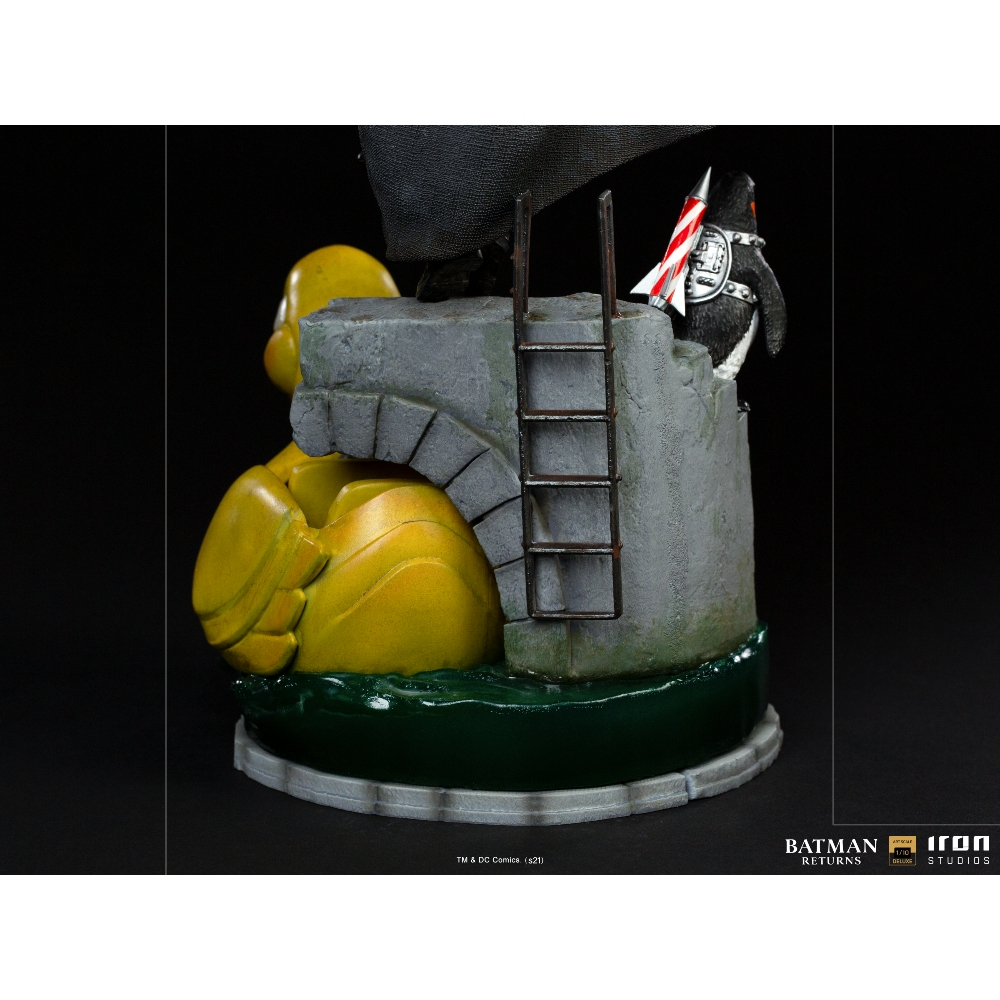 Statua di Batman Returns. Un personaggio pinguino con un razzo, che sale una scala su una struttura in pietra. Base gialla e verde.