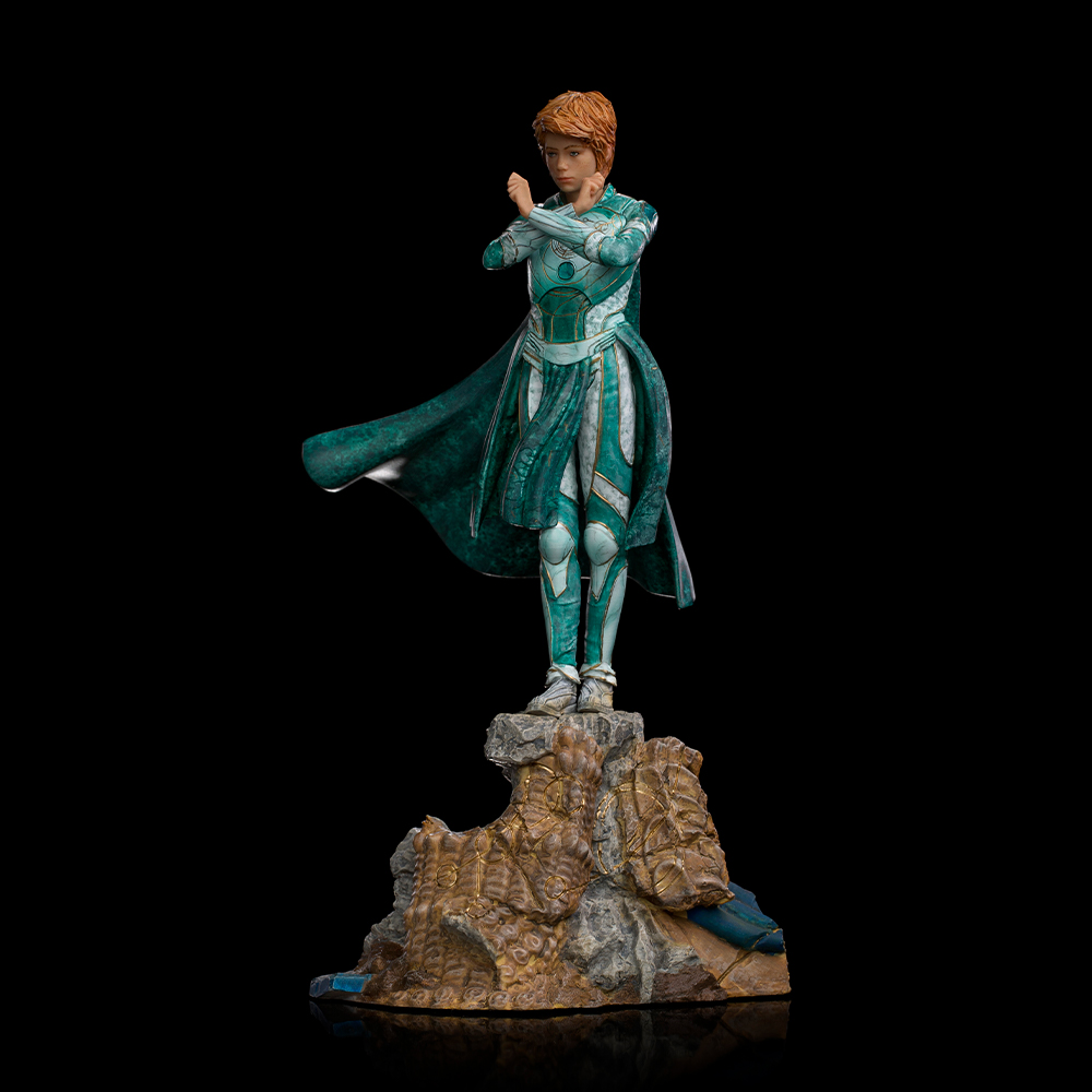 Una statua di una persona in un abito verde acqua e bianco è in piedi su una roccia contro uno sfondo nero. Ha i capelli rossi e le braccia incrociate.
