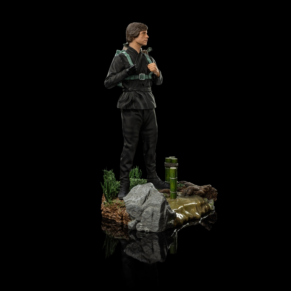 Eine Figur eines Mannes in Schwarz steht auf einer kleinen Plattform. Er hat einen grünen Rucksack. Die Plattform hat Felsen, Gras und Bambus.