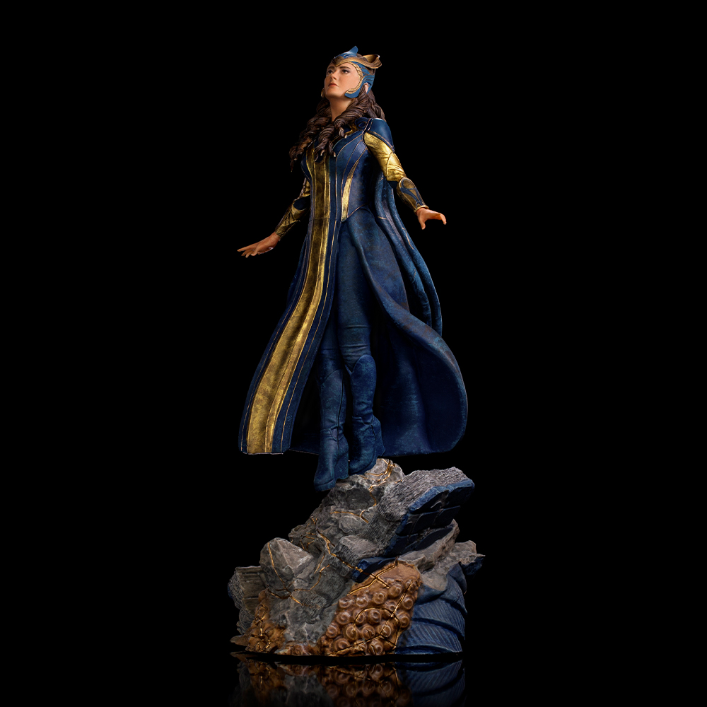 Statua di donna in abito blu e oro su base rocciosa, sfondo nero, guarda in alto con le braccia aperte.