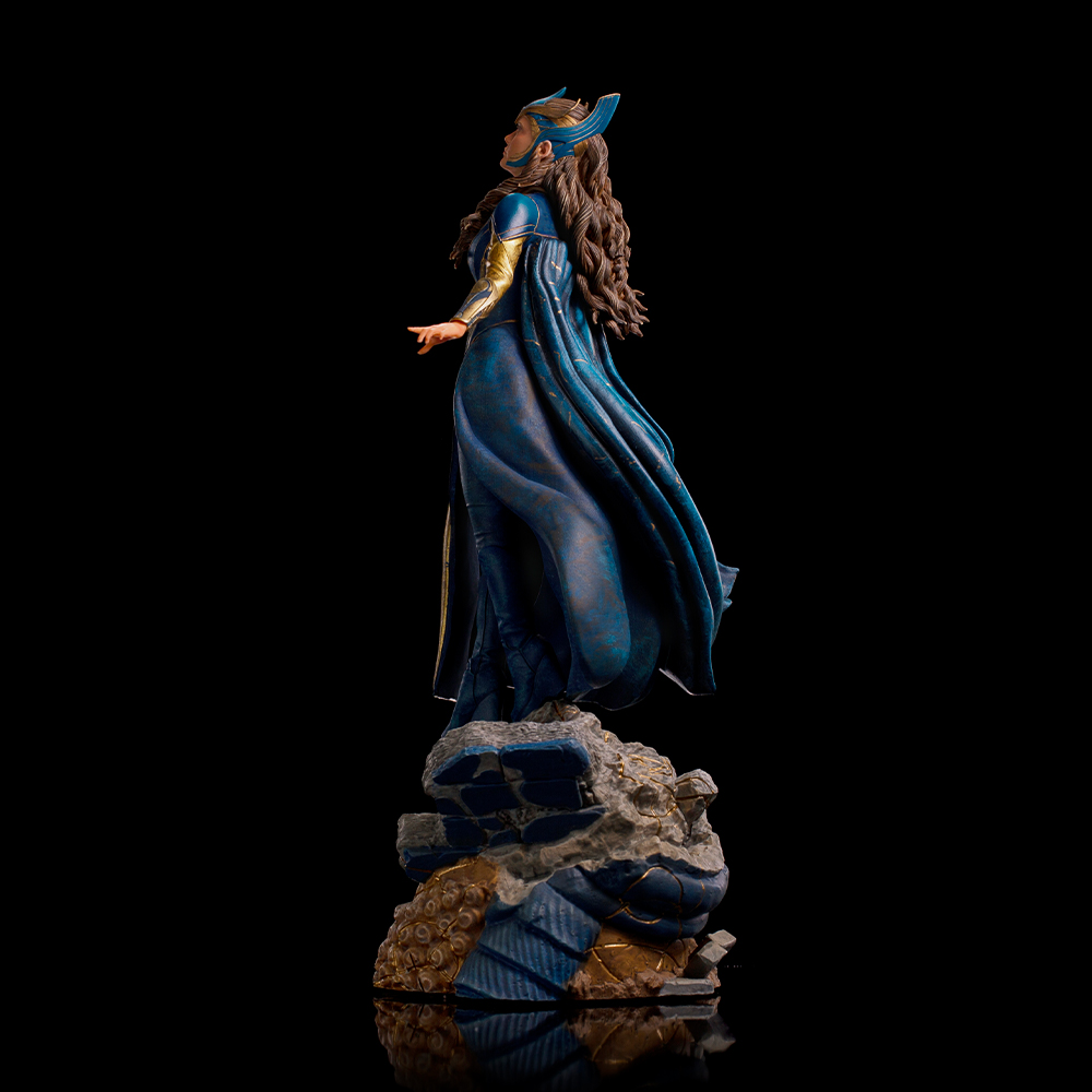 Una statua di una donna in abito e mantello blu, con dettagli dorati e un copricapo, in piedi su una roccia su sfondo nero.