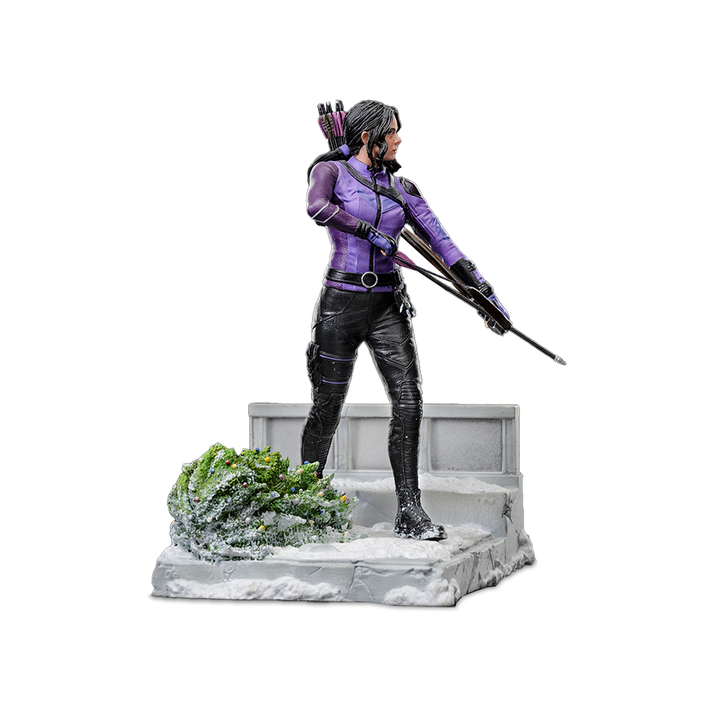 Statua di una donna in viola e nero, con arco e freccia. È su una piattaforma innevata con un piccolo cespuglio verde.