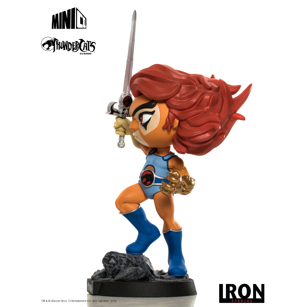Una statuina di Lion-O che tiene una spada. Indossa un completo blu, stivali e ha i capelli rossi. La statuina è su una base di roccia.