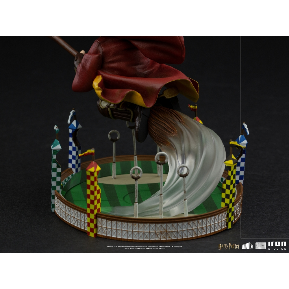Una figura di un mago su una scopa che vola sopra un campo di Quidditch, con cerchi e bandiere. La figura indossa un mantello rosso.