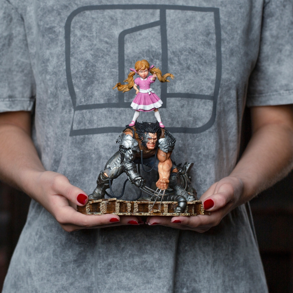 Una persona tiene in mano una statuetta di Wolverine con una bambina sopra. La bambina indossa un vestito rosa. La persona indossa una maglietta grigia.