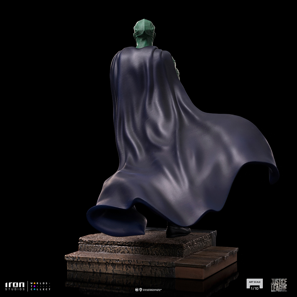 Statua di un supereroe dalla pelle verde con un mantello blu scuro, in piedi su una piattaforma di pietra, su sfondo nero.