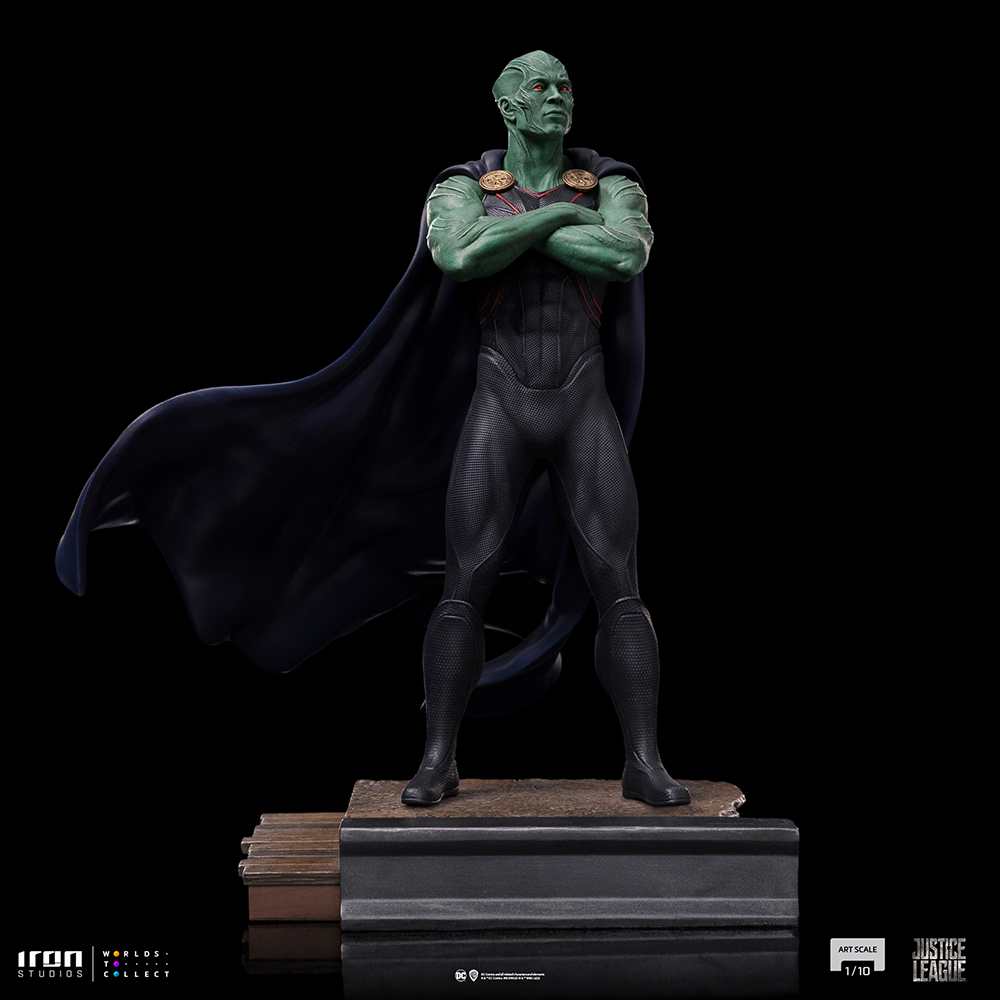 Statua di Martian Manhunter, pelle verde, tuta nera, mantello blu, braccia conserte. In piedi su una piattaforma con sfondo scuro.