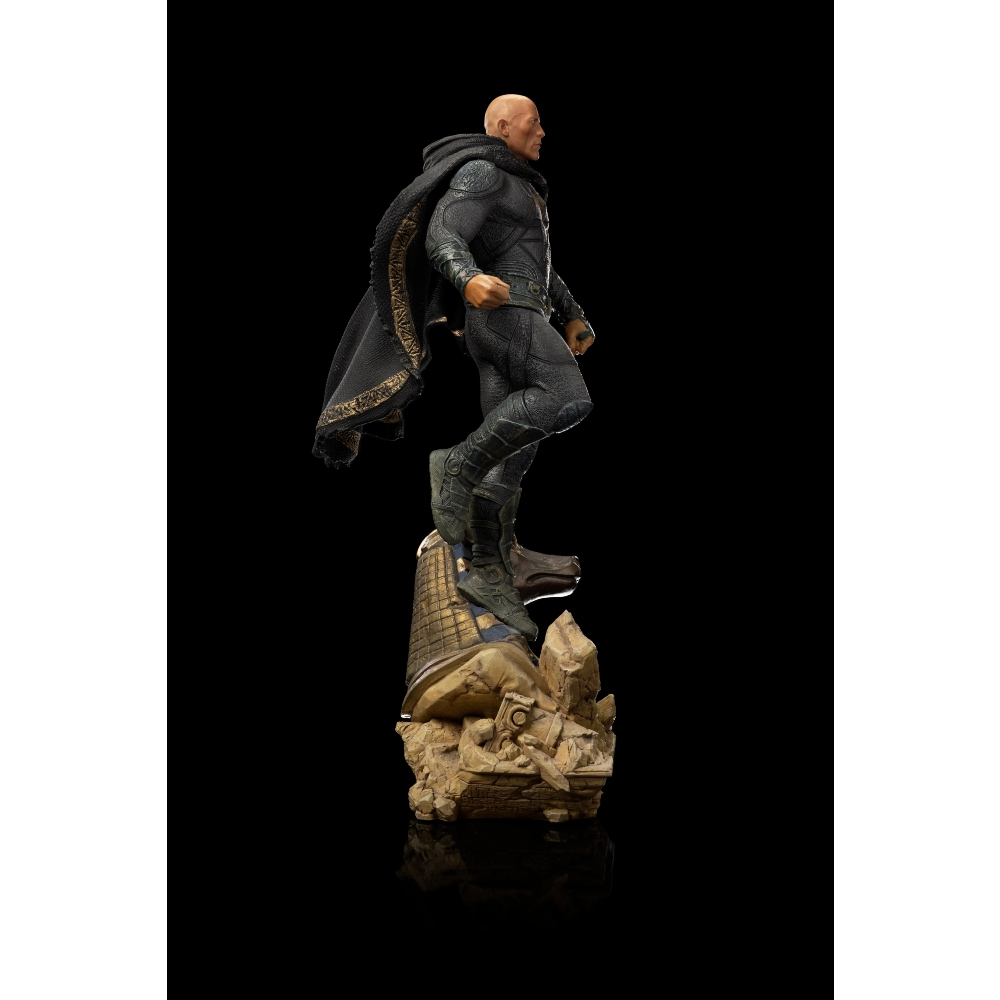 Una figura calva in costume nero con un mantello su una base di pietra. Lo sfondo è nero.