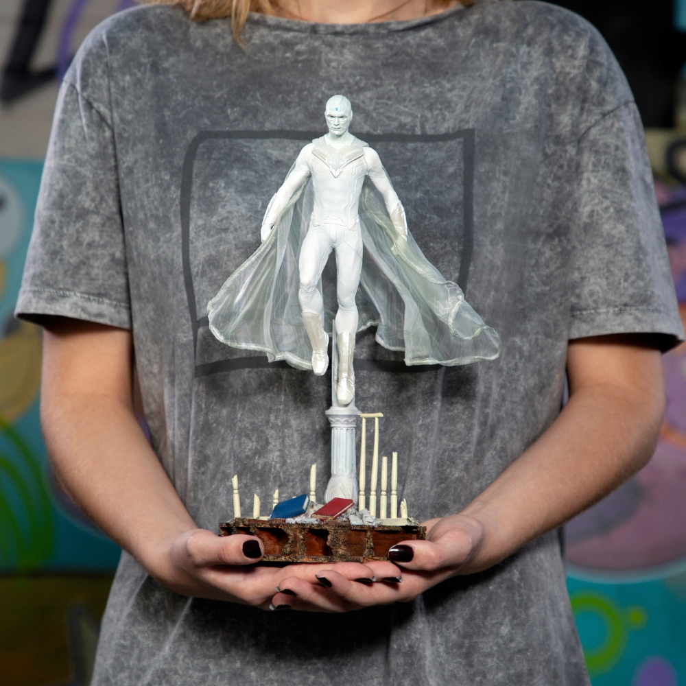 Una persona tiene in mano una statuetta bianca di Vision. Si trova su un pilastro sopra una base con piccoli libri e bastoncini. La persona indossa una maglietta grigia.