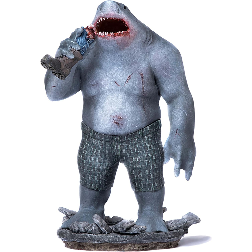 Una statua di un ibrido uomo-squalo, che tiene e mangia una persona. La figura indossa pantaloncini e si trova su delle rocce.