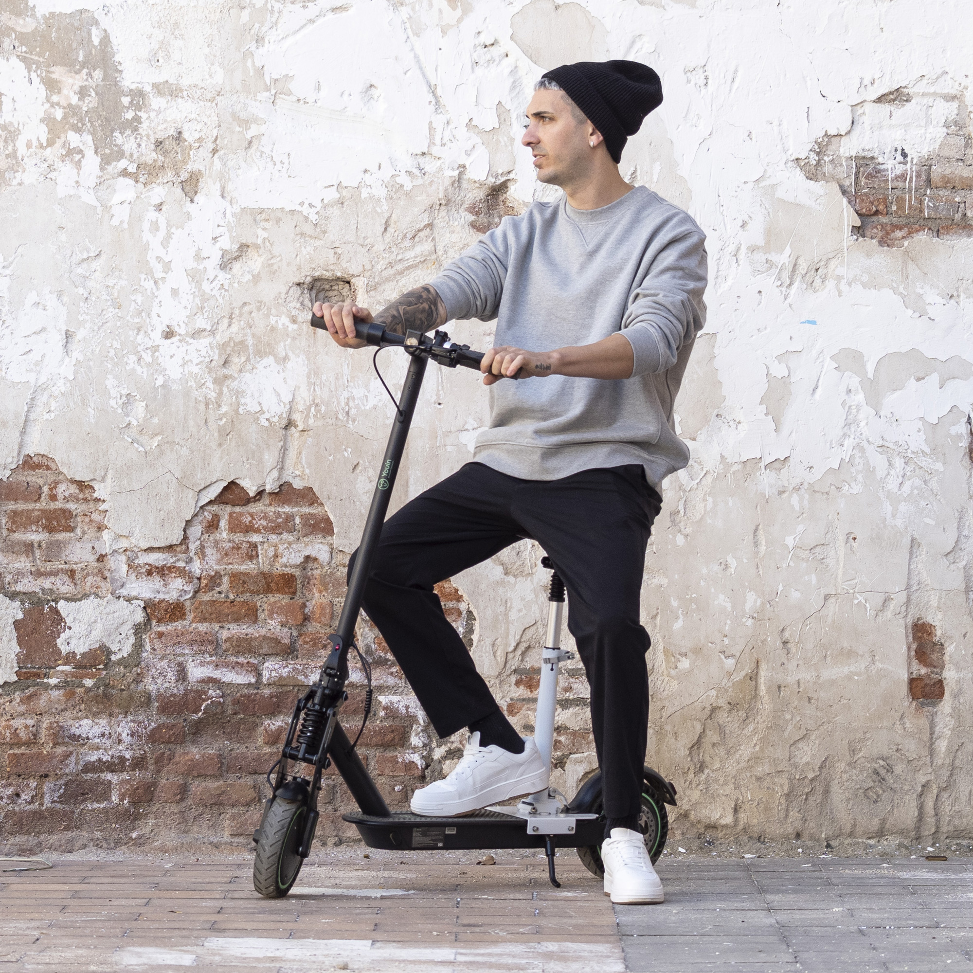 Hombre en patinete, con gorro negro, sudadera gris y pantalones negros. Fondo de pared de ladrillos.