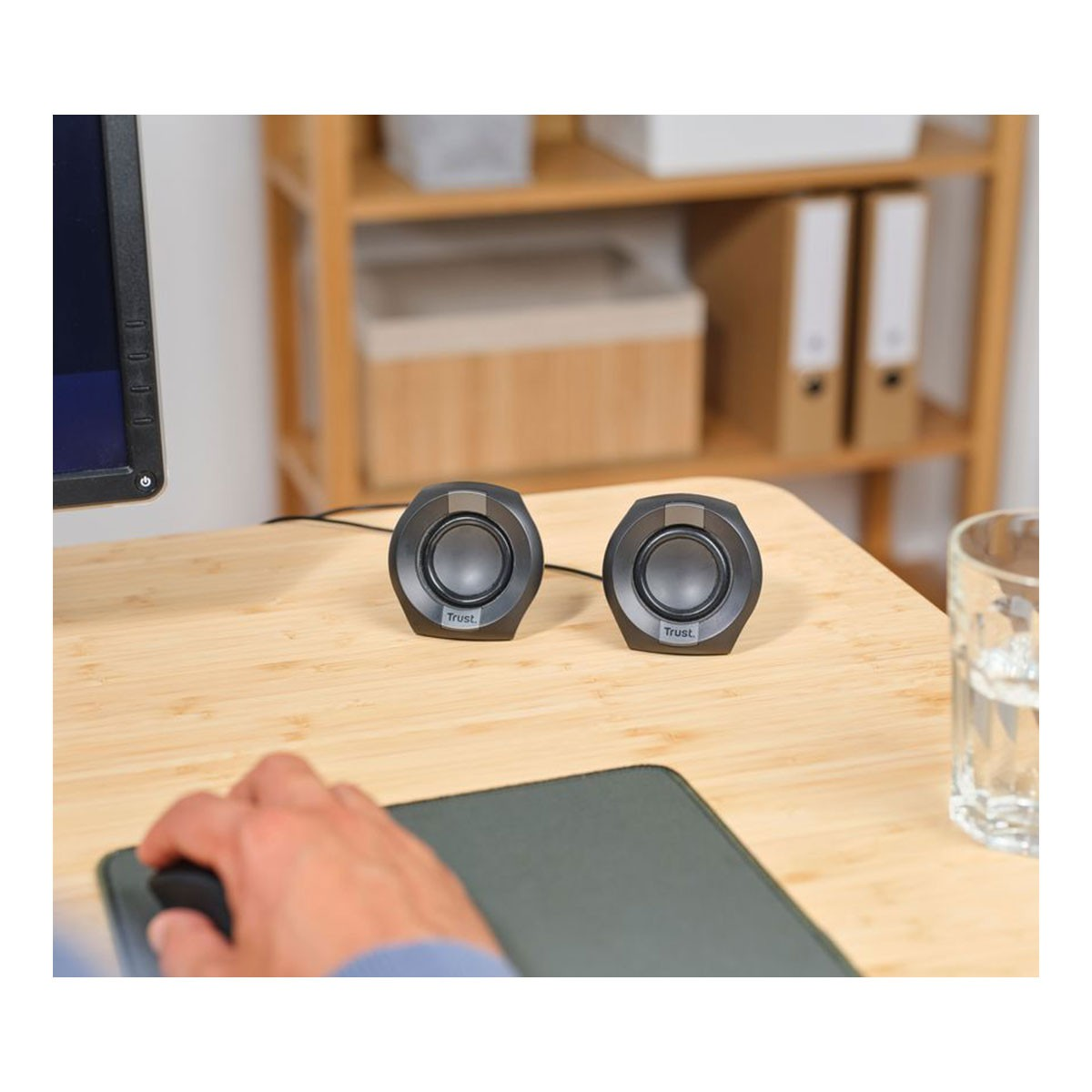 Dwa czarne głośniki z napisem 'Trust' na drewnianym biurku. Ręka osoby na myszy i szklanka wody po prawej stronie.