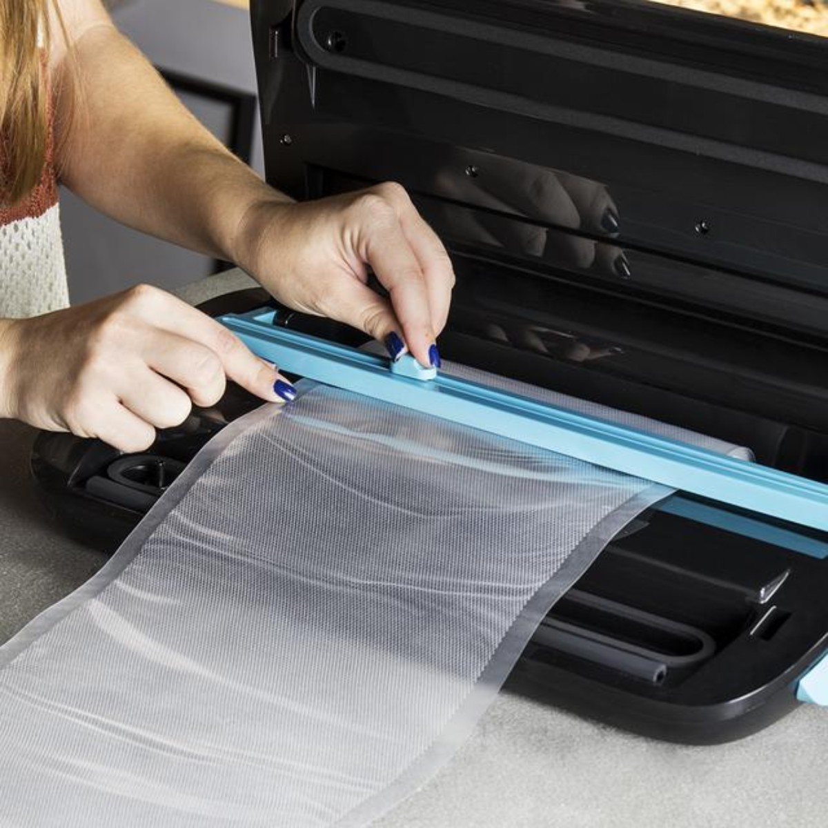 Osoba używa zgrzewarki próżniowej. Ręce umieszczają plastikową torbę w maszynie. Maszyna jest czarno-niebieska.