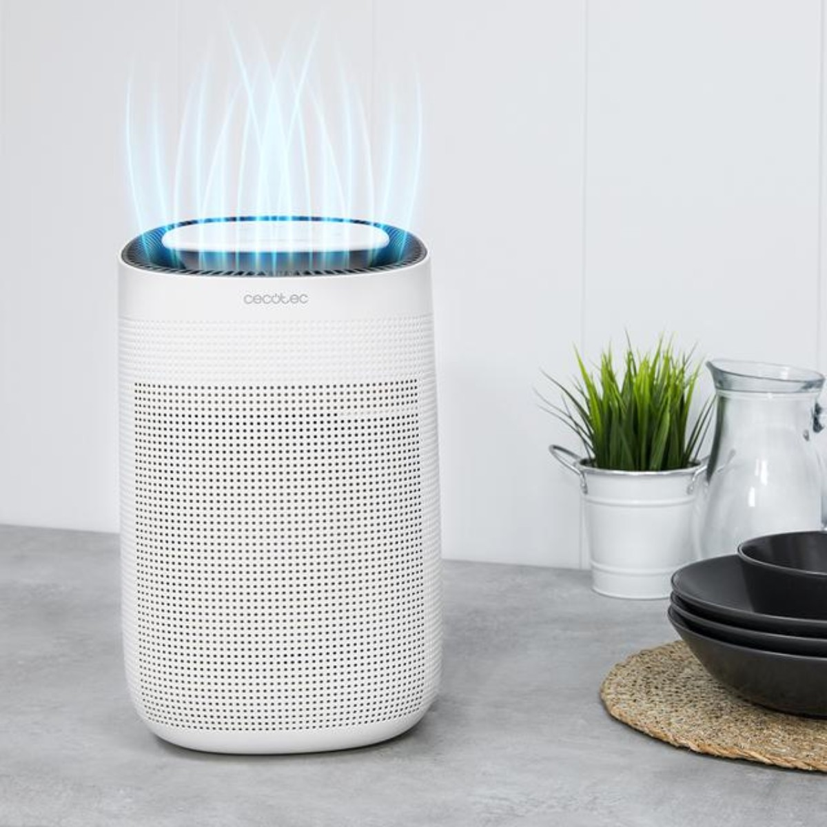 Biały oczyszczacz powietrza z efektem niebieskiego światła. Roślina, miski i dzbanek na szarym blacie. Biała ściana w tle.