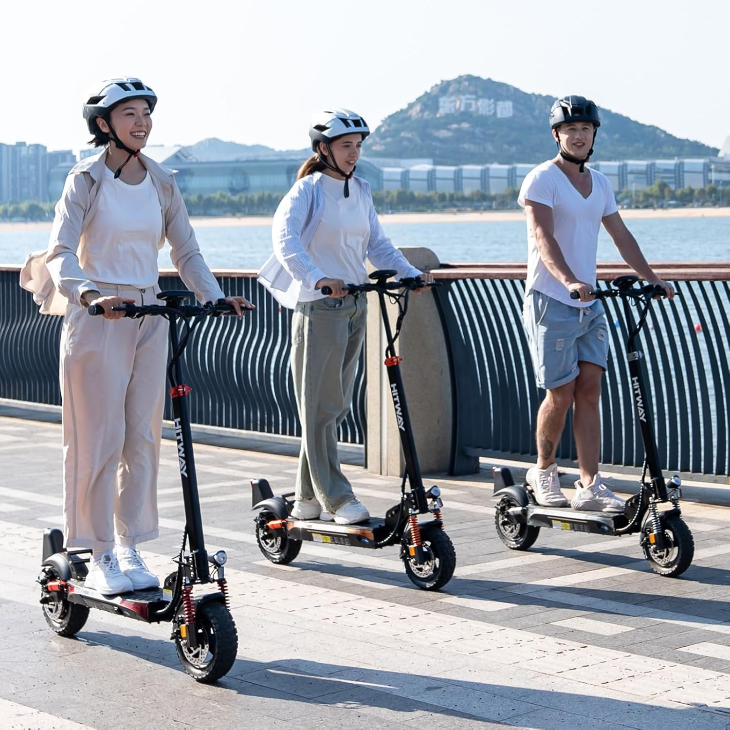 Trzy osoby jeżdżą na hulajnogach elektrycznych w słoneczny dzień. Mają kaski. W tle widać górę, budynki i morze.