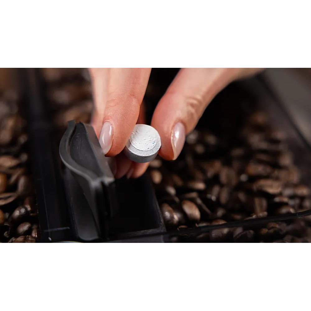 Una mano tiene in mano una piccola compressa rotonda argentata sopra un contenitore di chicchi di caffè, un componente di plastica nera è in vista.