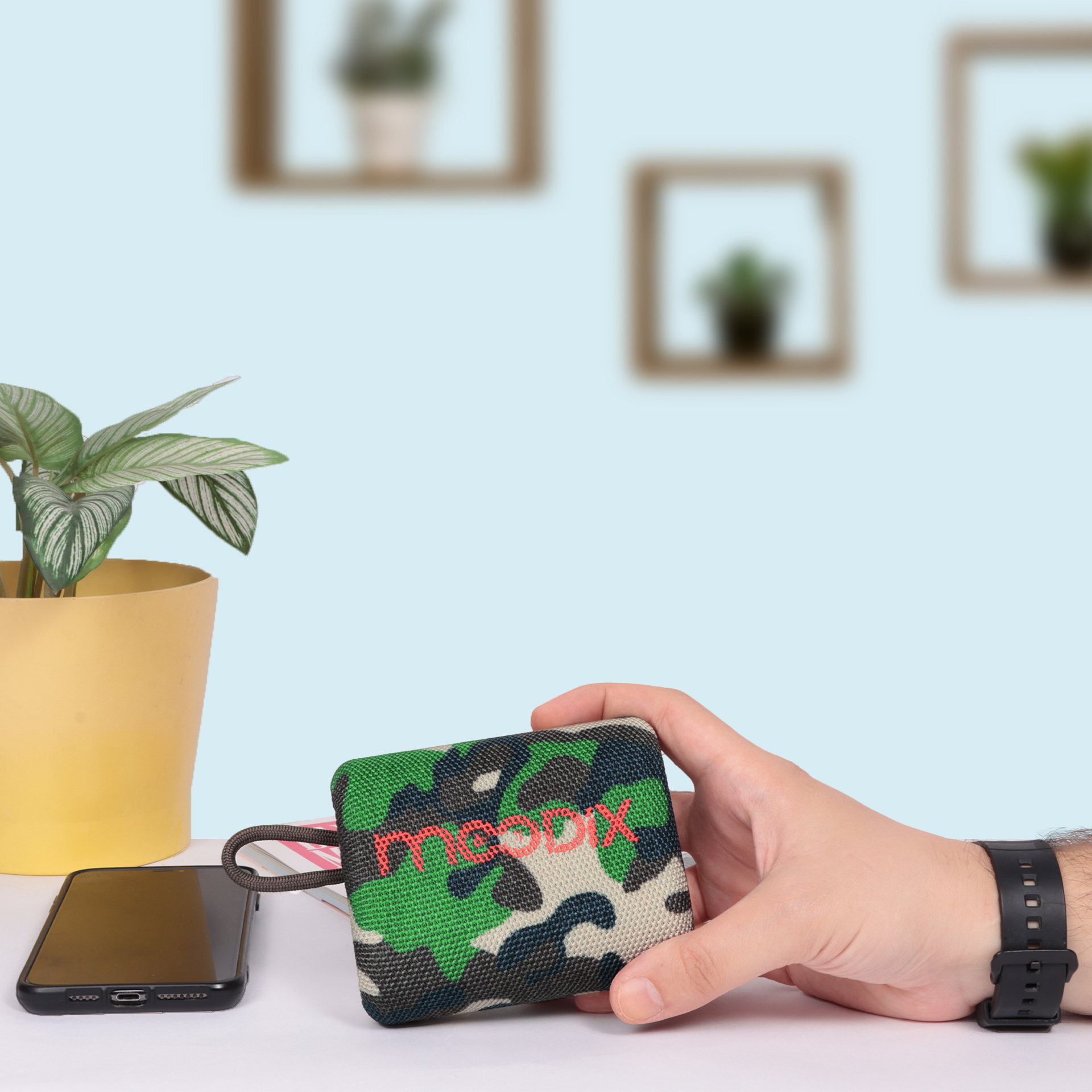 El, kırmızı yazılı yeşil kamuflaj hoparlör tutuyor. Arka planda bir telefon, bitki ve duvar dekoru var.