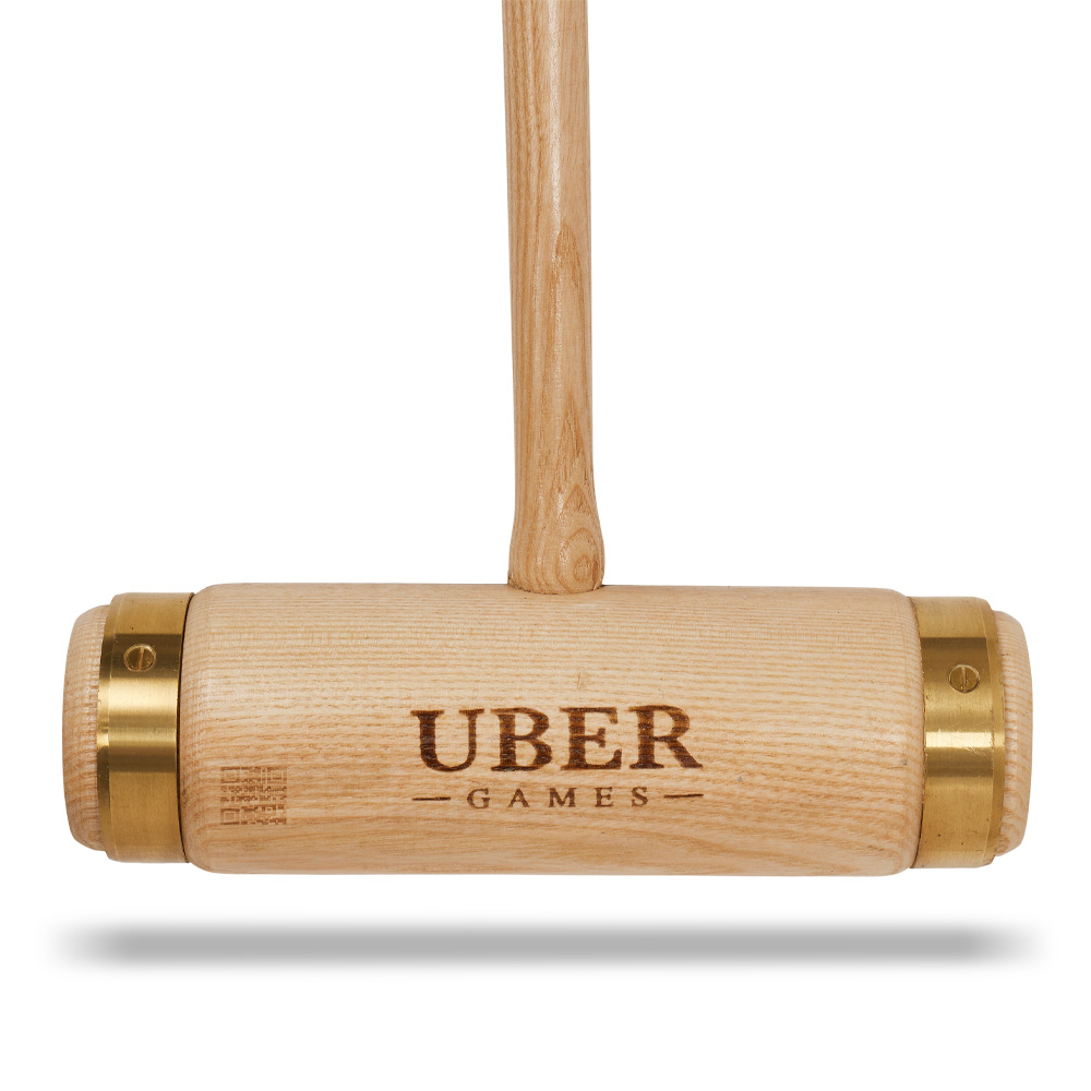 Ein hölzerner Krocket-Schläger mit Messingbeschlägen und dem Text 'UBER GAMES' an der Seite. Er befindet sich auf weißem Hintergrund.